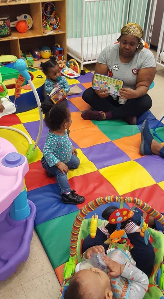 Person reading a book to two children, with a baby in a bouncer nearby
