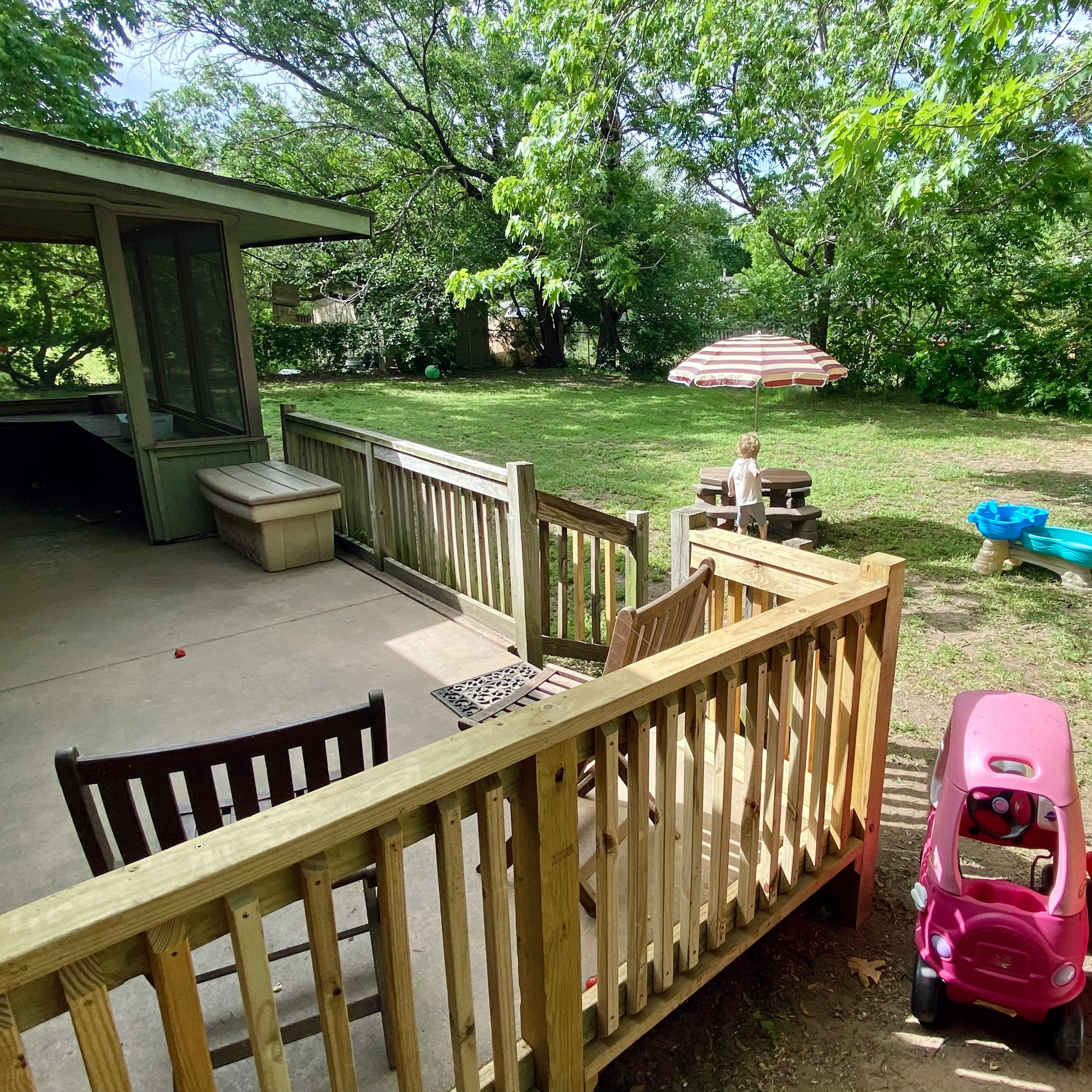 Patio with wooden railing overlooking a grassy yard with a child near a picnic table