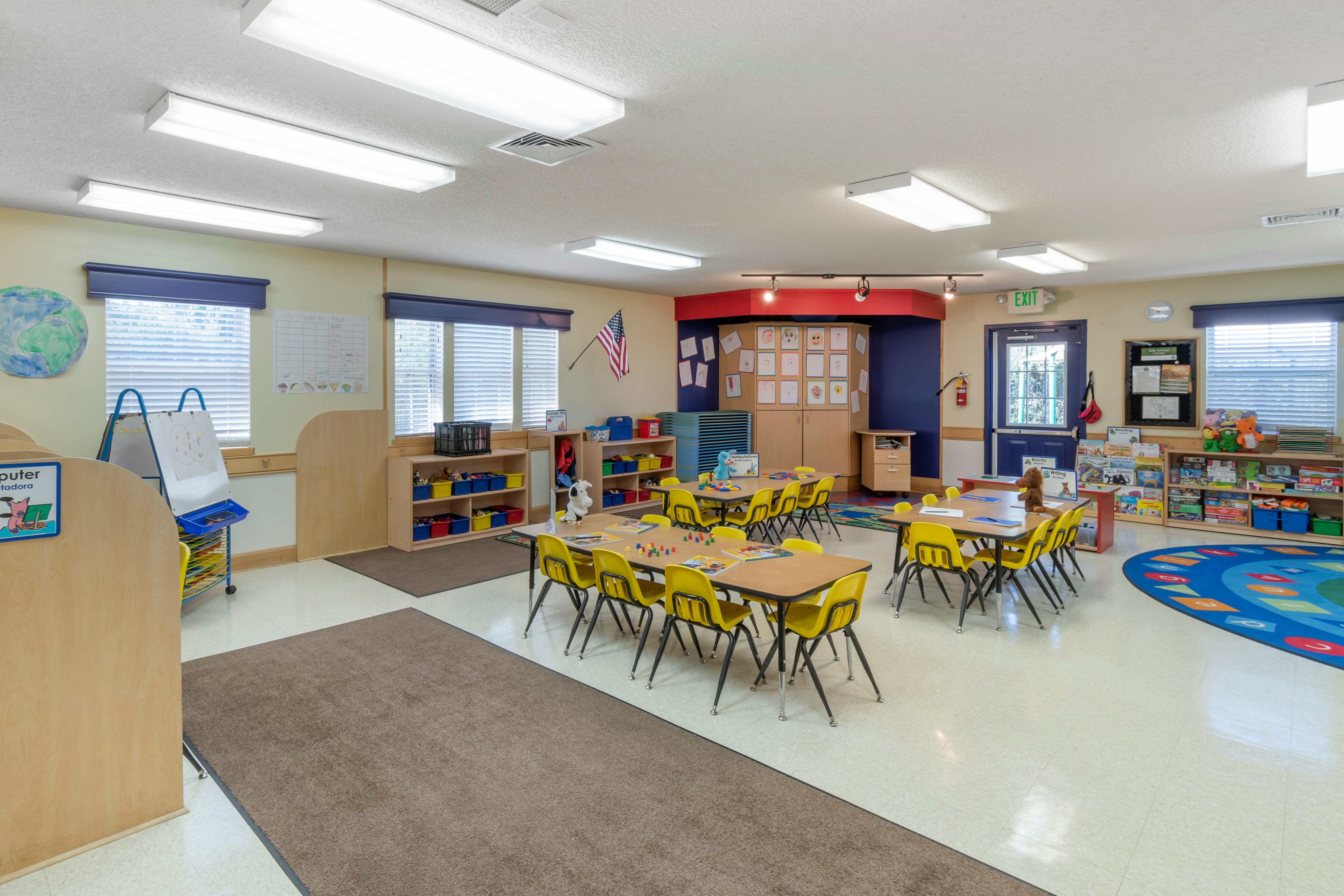 Classroom with small tables and chairs, shelves with toys, and an alphabet rug