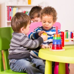 Two children playing with toys at a table, another child in background