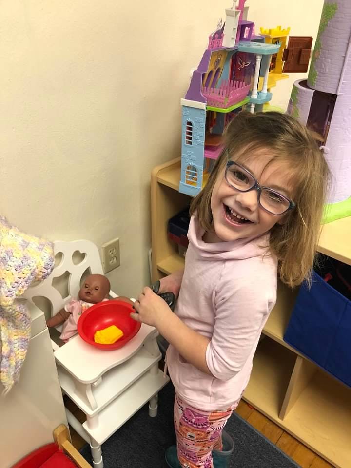 Child in glasses smiling, standing next to a doll in a high chair