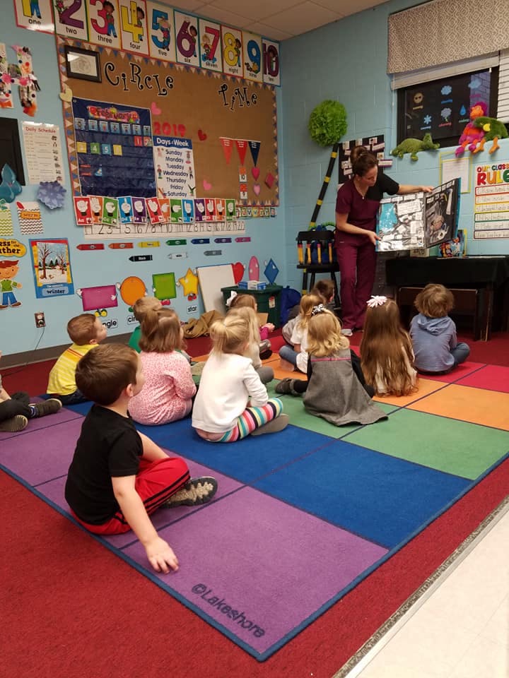 Adult reading a book to children sitting on a colorful rug in a classroom