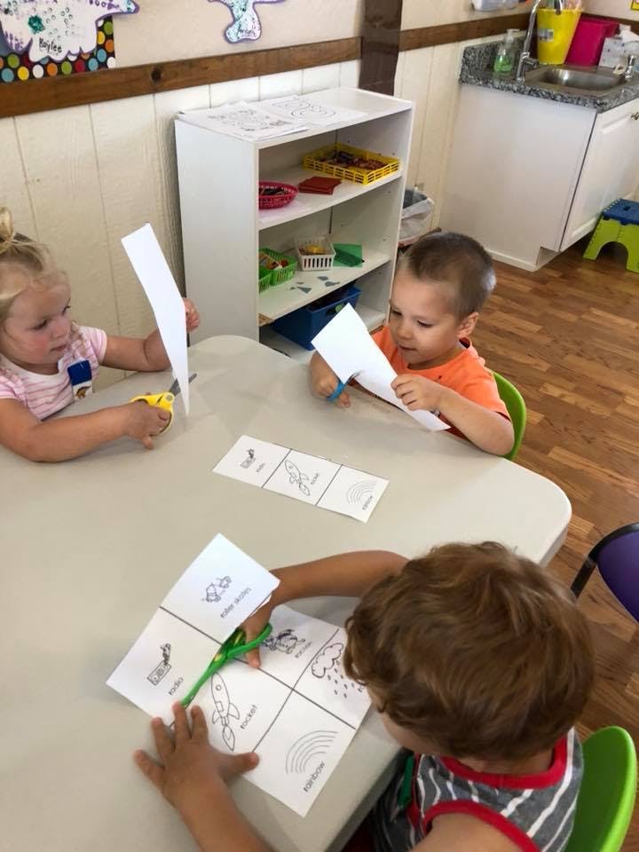 Three children cutting paper at a table in a classroom