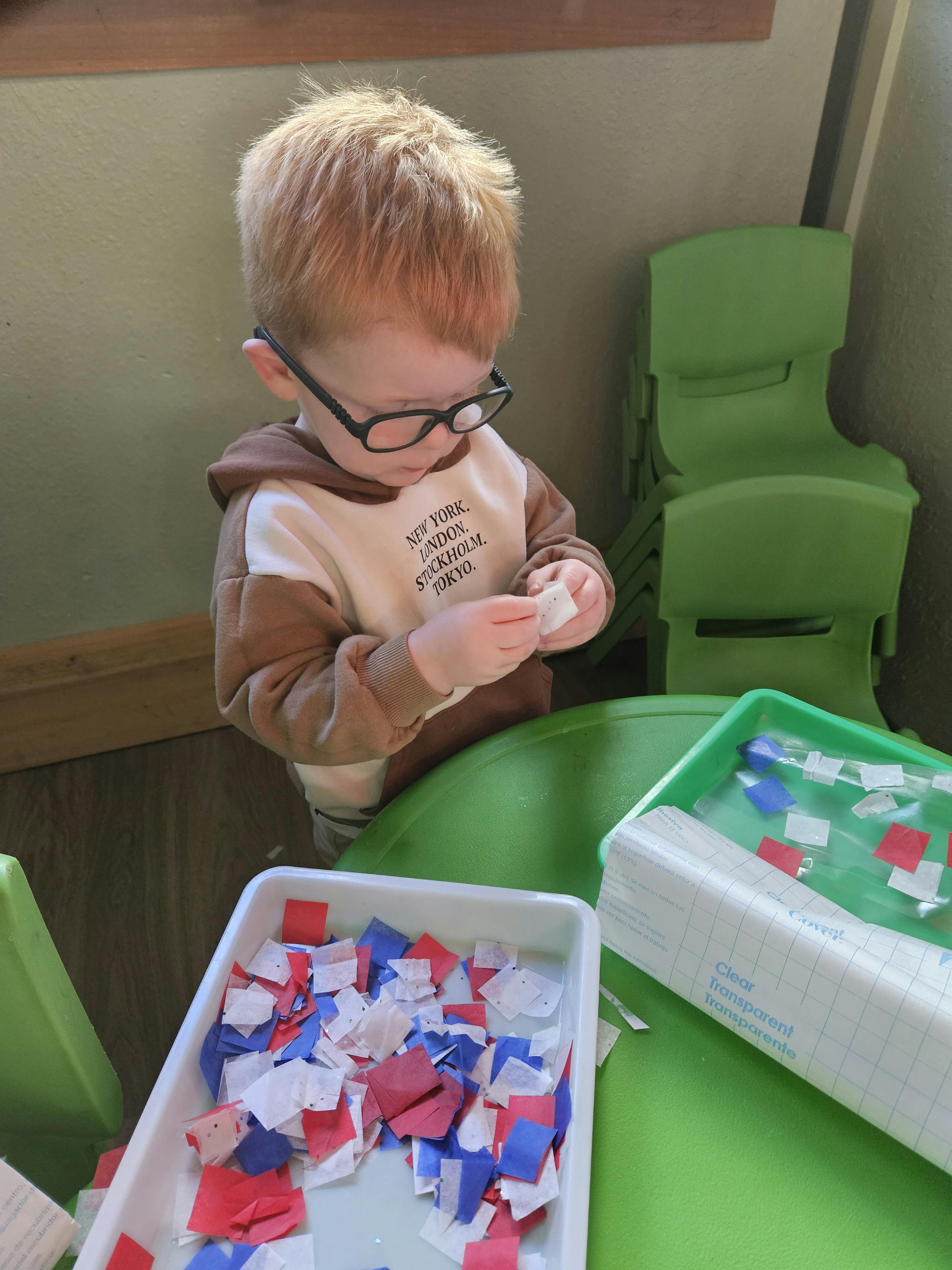 Child wearing glasses holding a small white object over a table with craft supplies