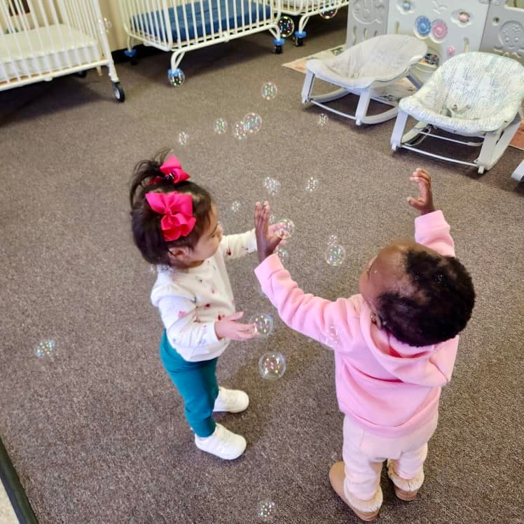 Two children reaching for bubbles in a room with cribs in the background