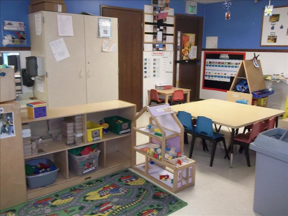 Children's classroom with tables, chairs, shelving with toys, and a dollhouse