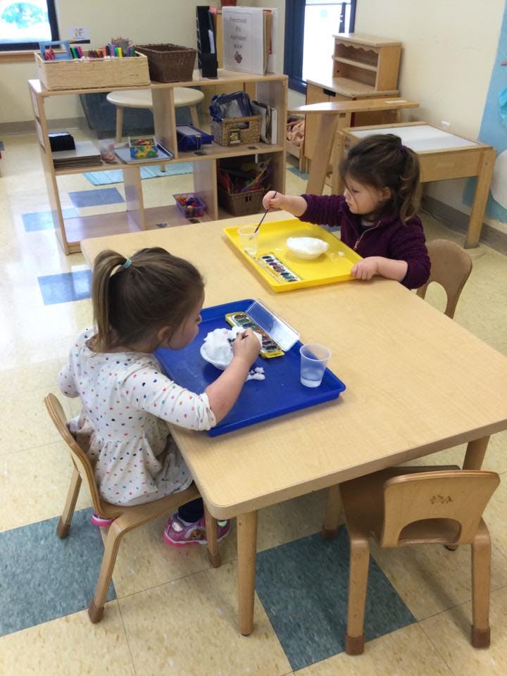 Two children at a table with trays, one painting and one working with white material