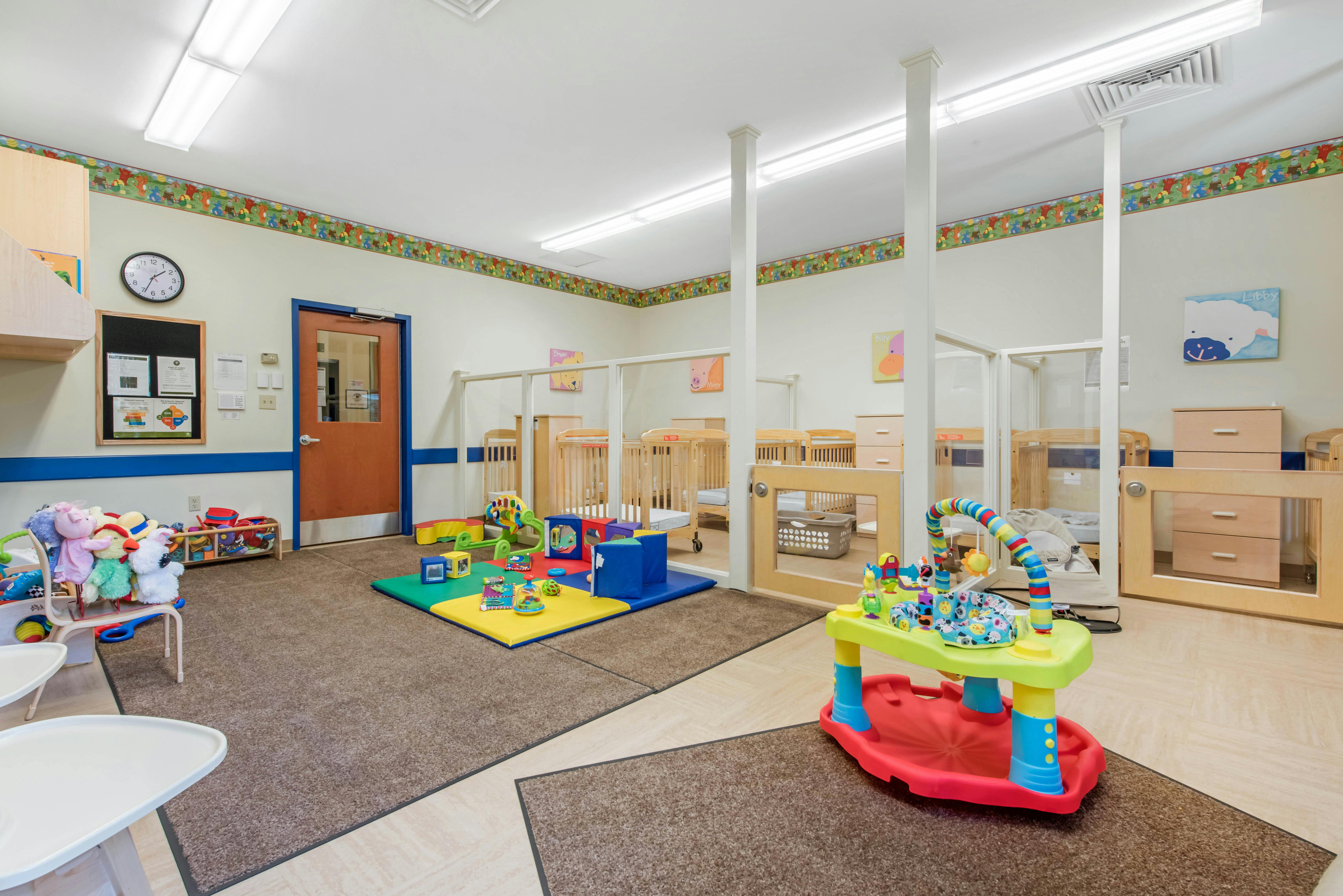 An indoor play area with colorful mats, toys, cribs behind a partition, and a door