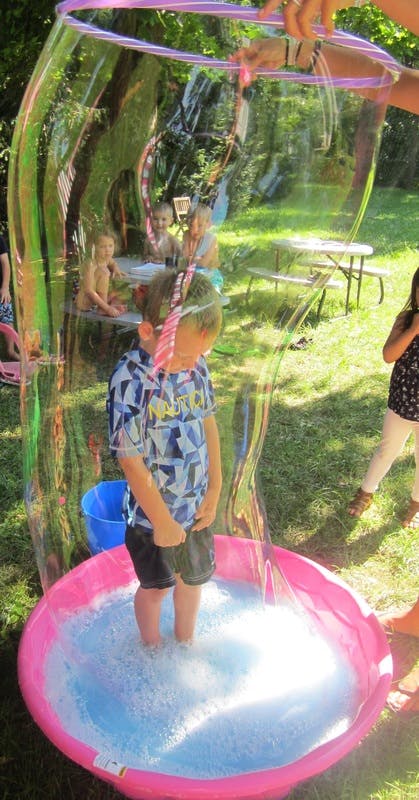 Child standing in a bubble solution in a pink pool with a large bubble forming around them