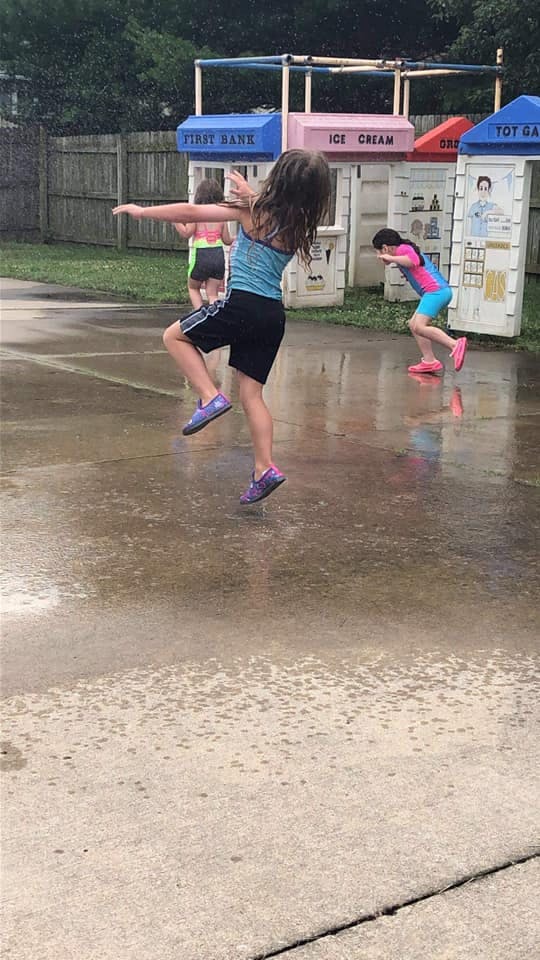 Child balancing on one leg on wet pavement, other children in background