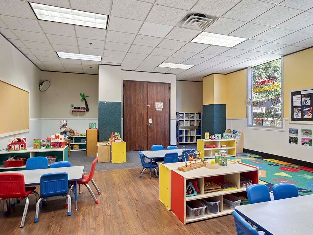 Empty classroom with small tables and chairs, shelves with toys, and colorful rugs