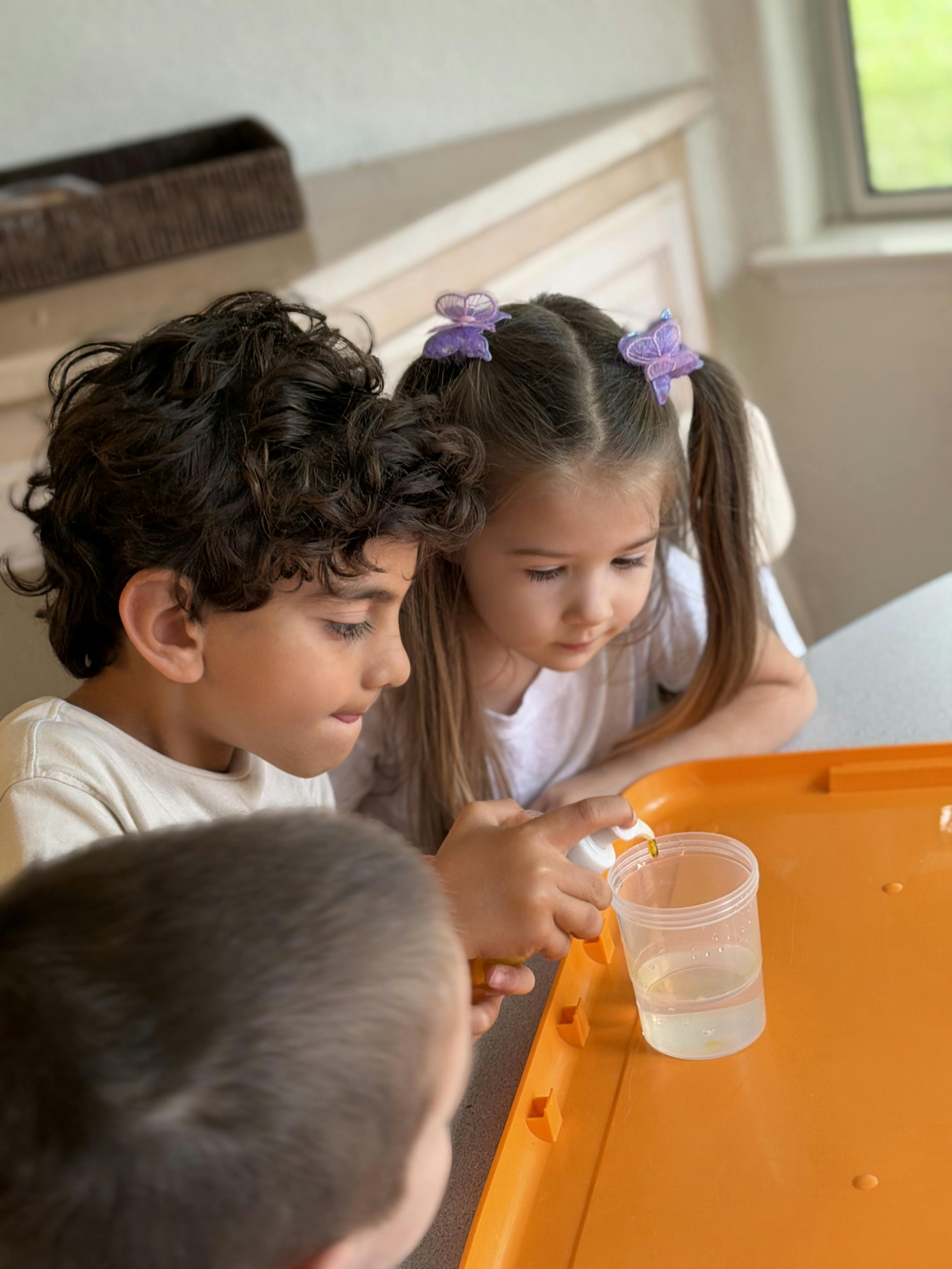 Children watching another child squeeze liquid from a pump bottle into a cup