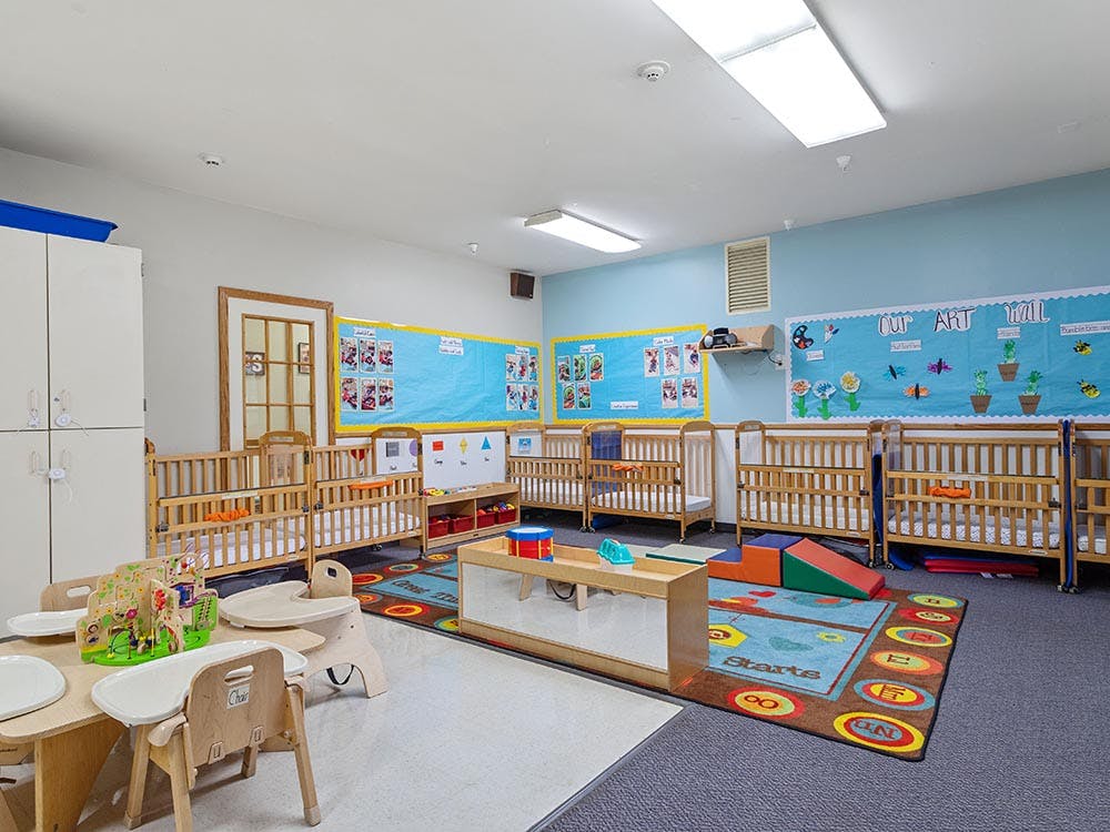 Cribs inside a brightly lit childcare room with infant play equipment