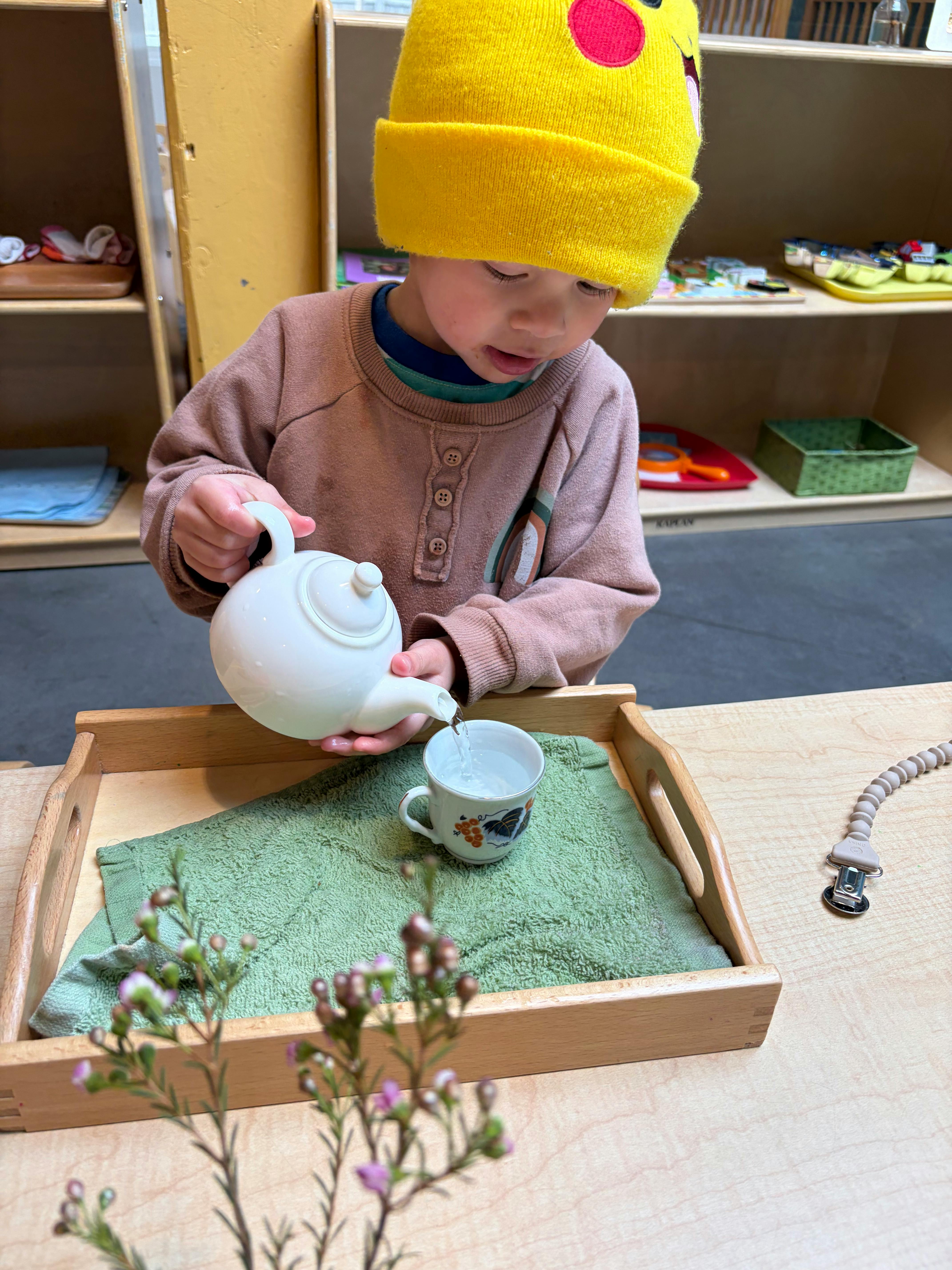 Child wearing a yellow hat pouring water from a teapot into a cup