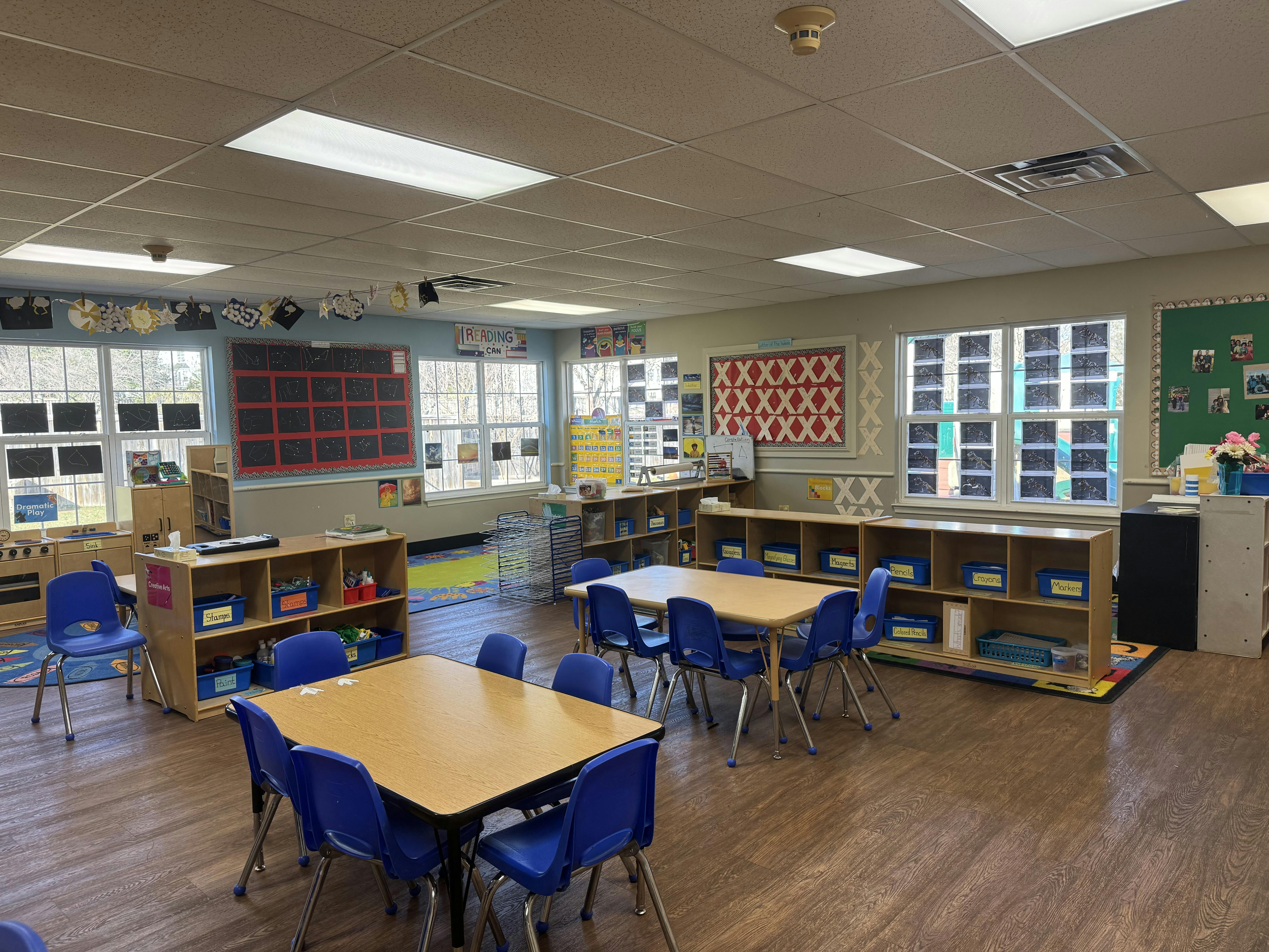 Classroom with small tables and chairs, shelves with bins, and windows