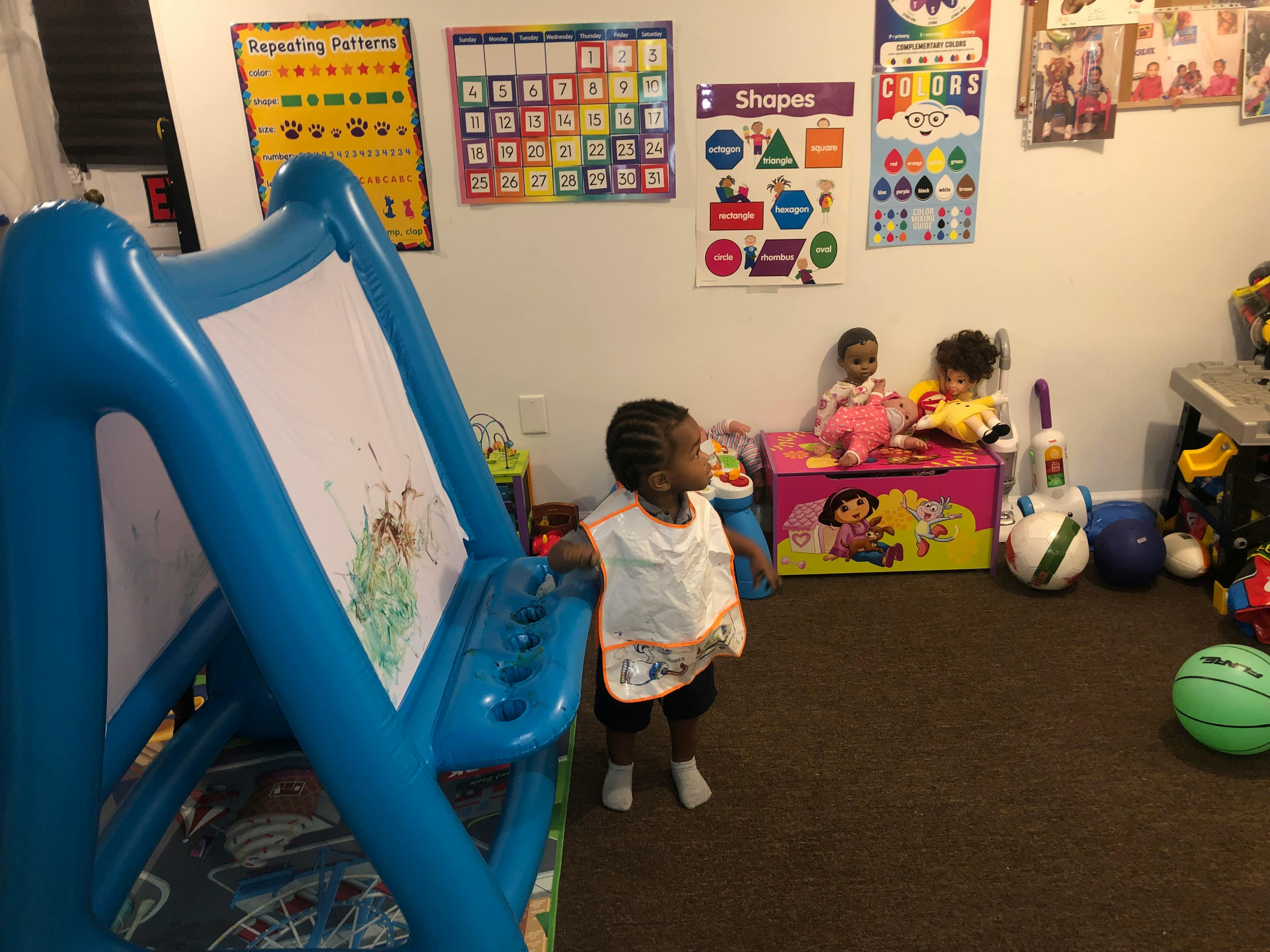 Child standing near an easel in a playroom with other children and toys