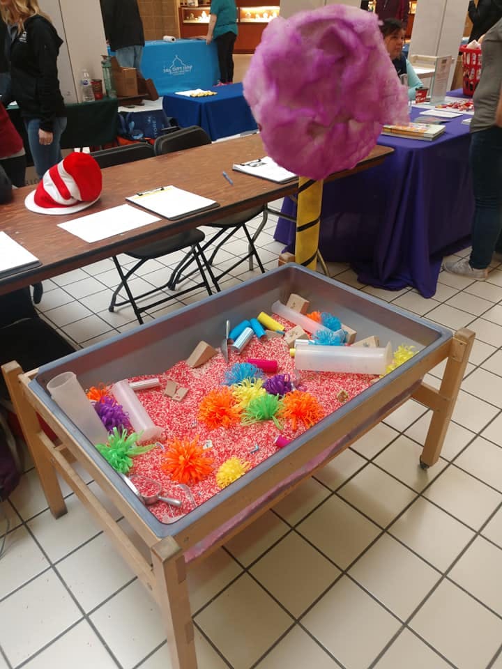 Sensory table filled with red rice, colorful pom-poms, and plastic tubes