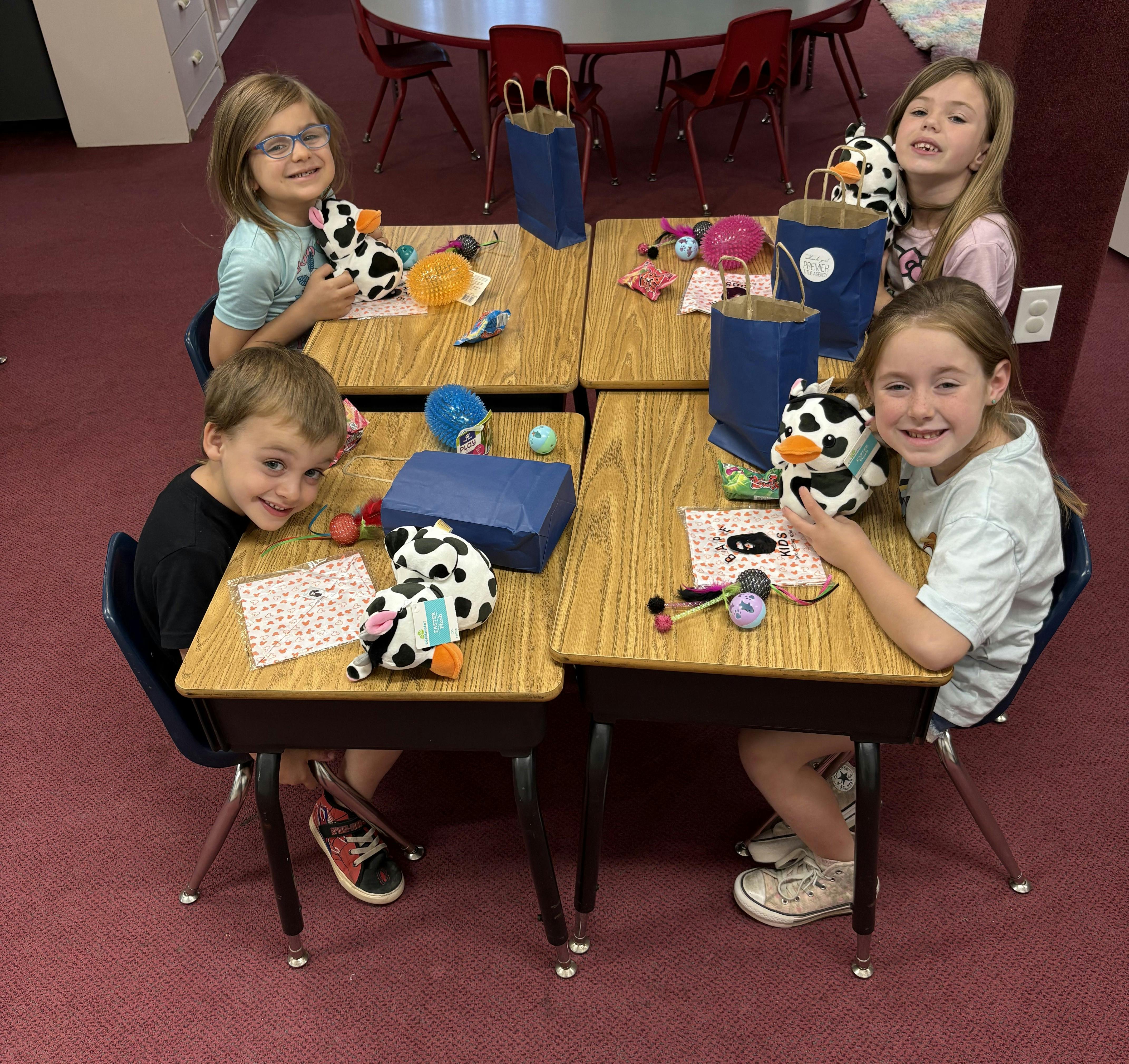 Four children sitting at desks with stuffed animals and craft supplies