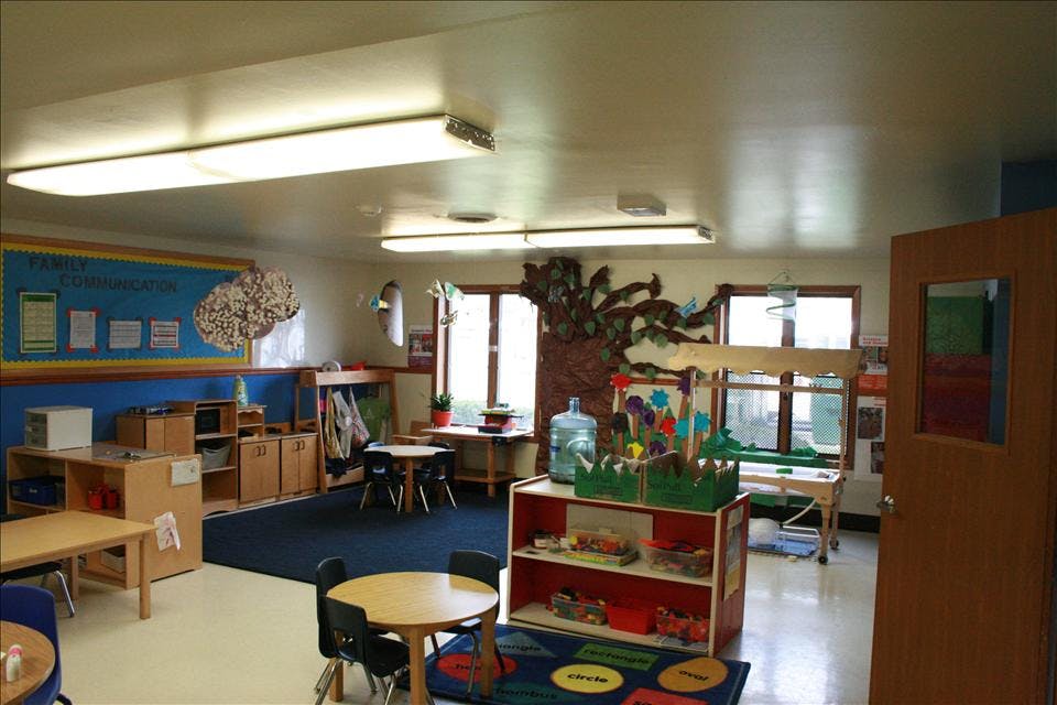 Classroom with small tables and chairs, shelves with toys, and bulletin board