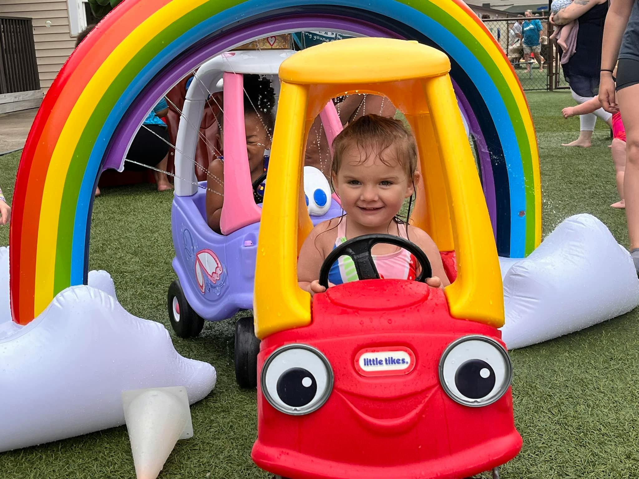 Child in a red toy car under a rainbow arch