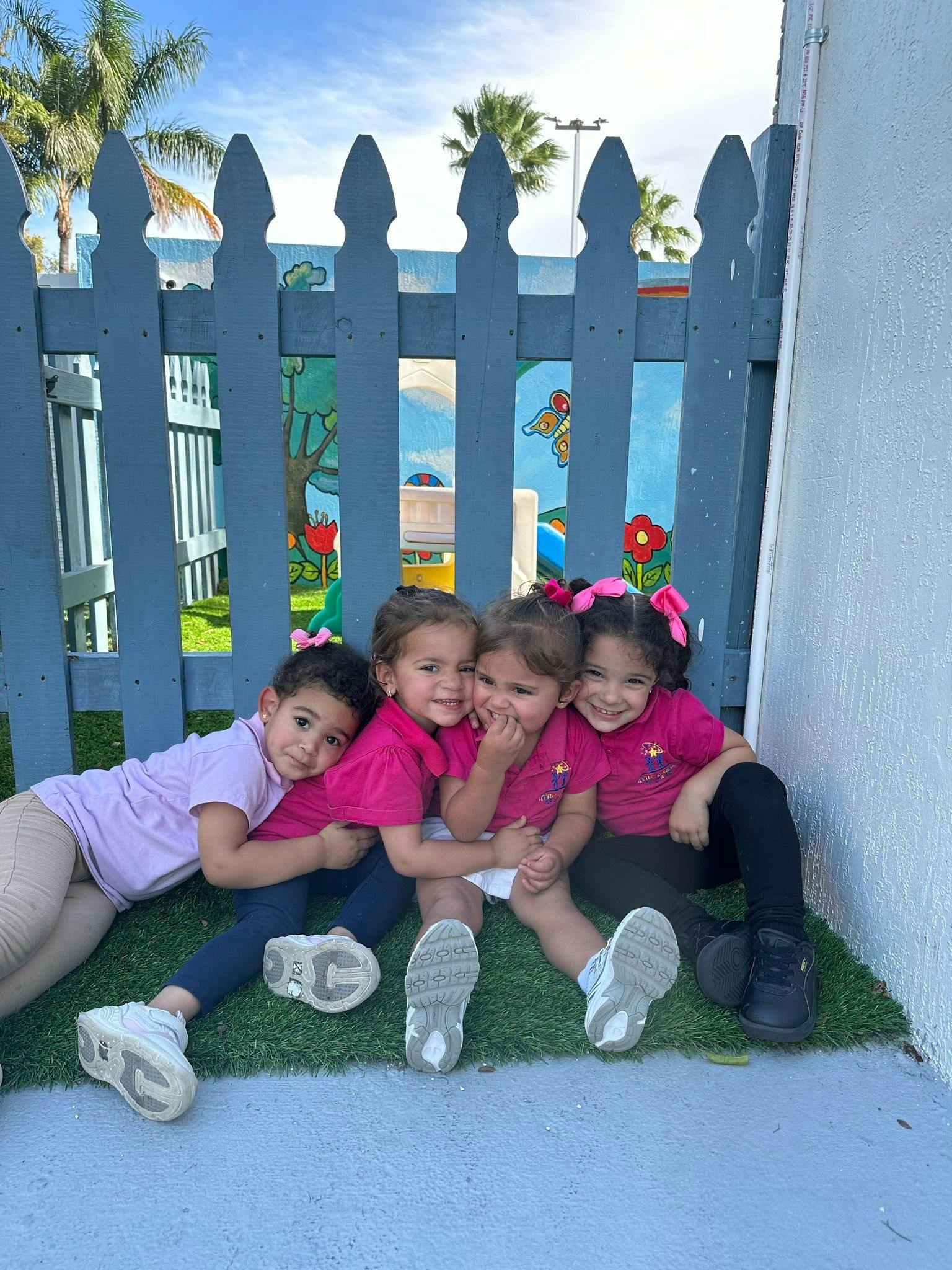Four children sitting on grass in front of a blue picket fence