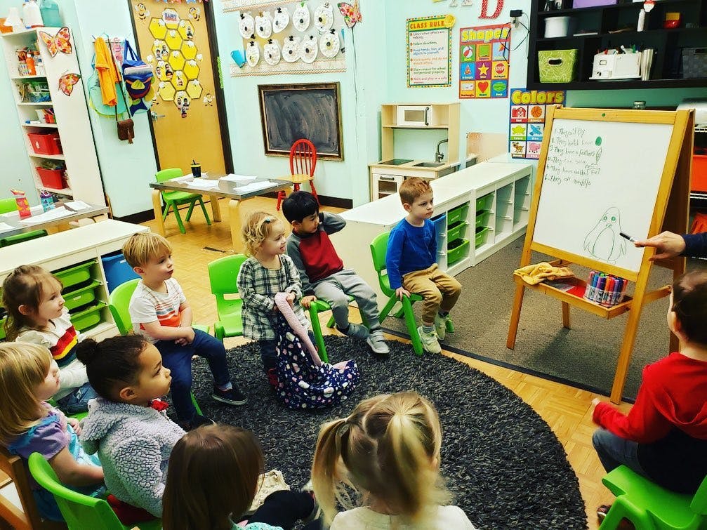 Children sitting on a rug and chairs watching a person point to a whiteboard