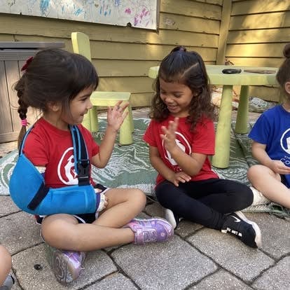 Two children sitting on the ground, one with a blue arm sling