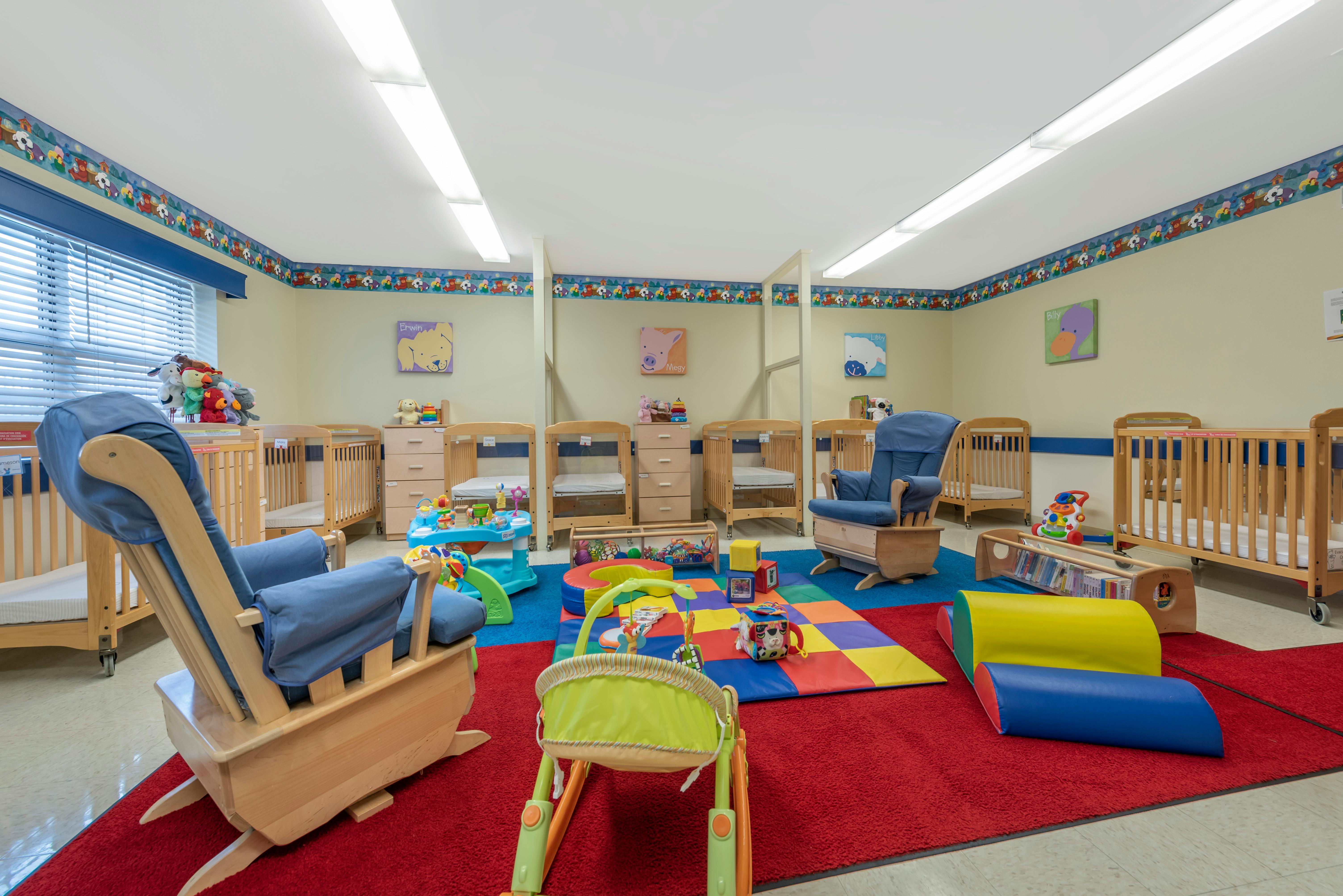 A colorful childcare room with cribs, rocking chairs, and play mats