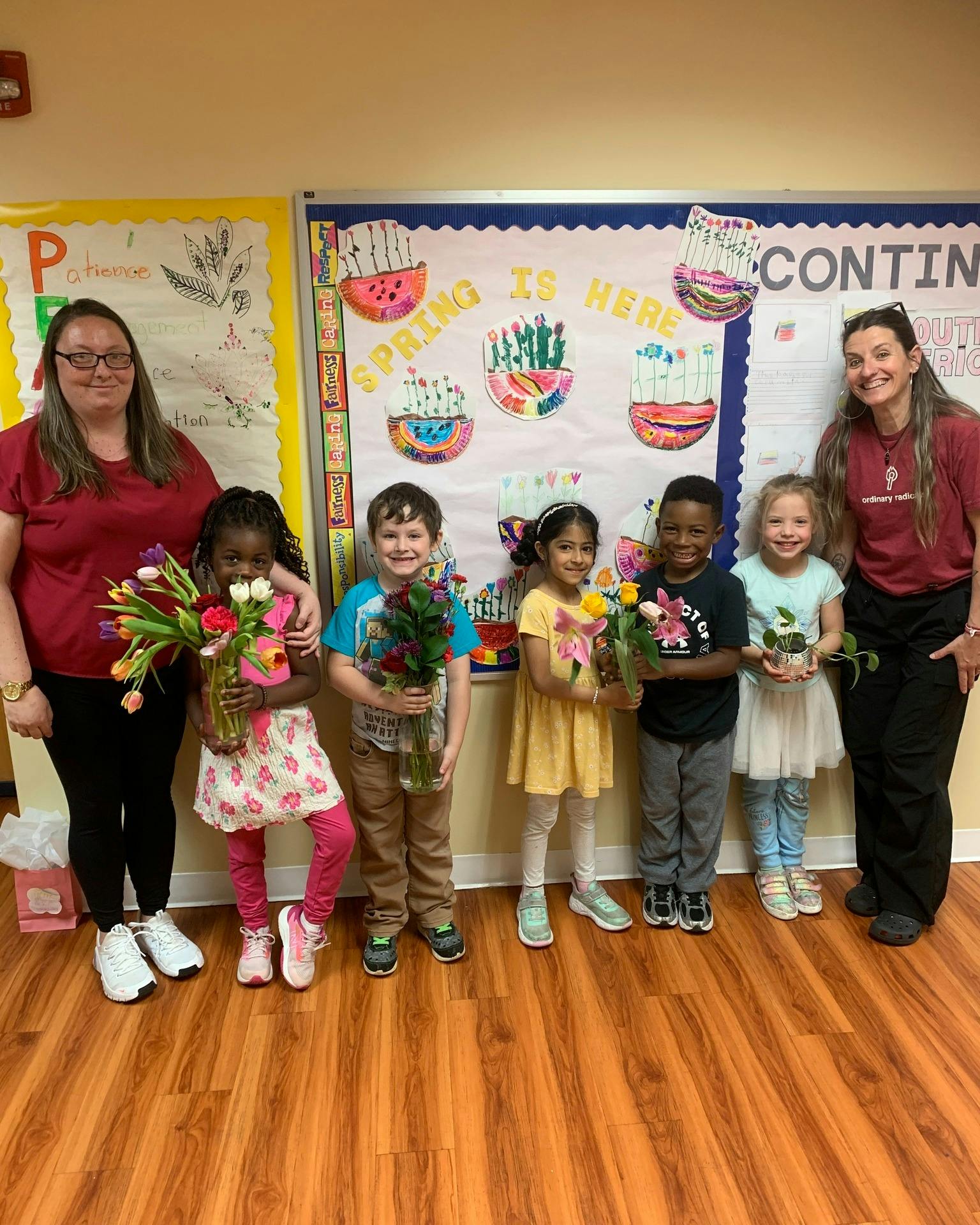 Two adults and five children holding flowers and plants in a classroom