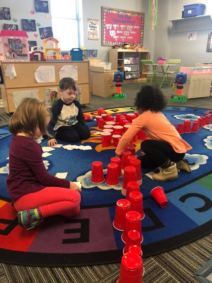 Three children playing with red cups on a colorful rug in a classroom