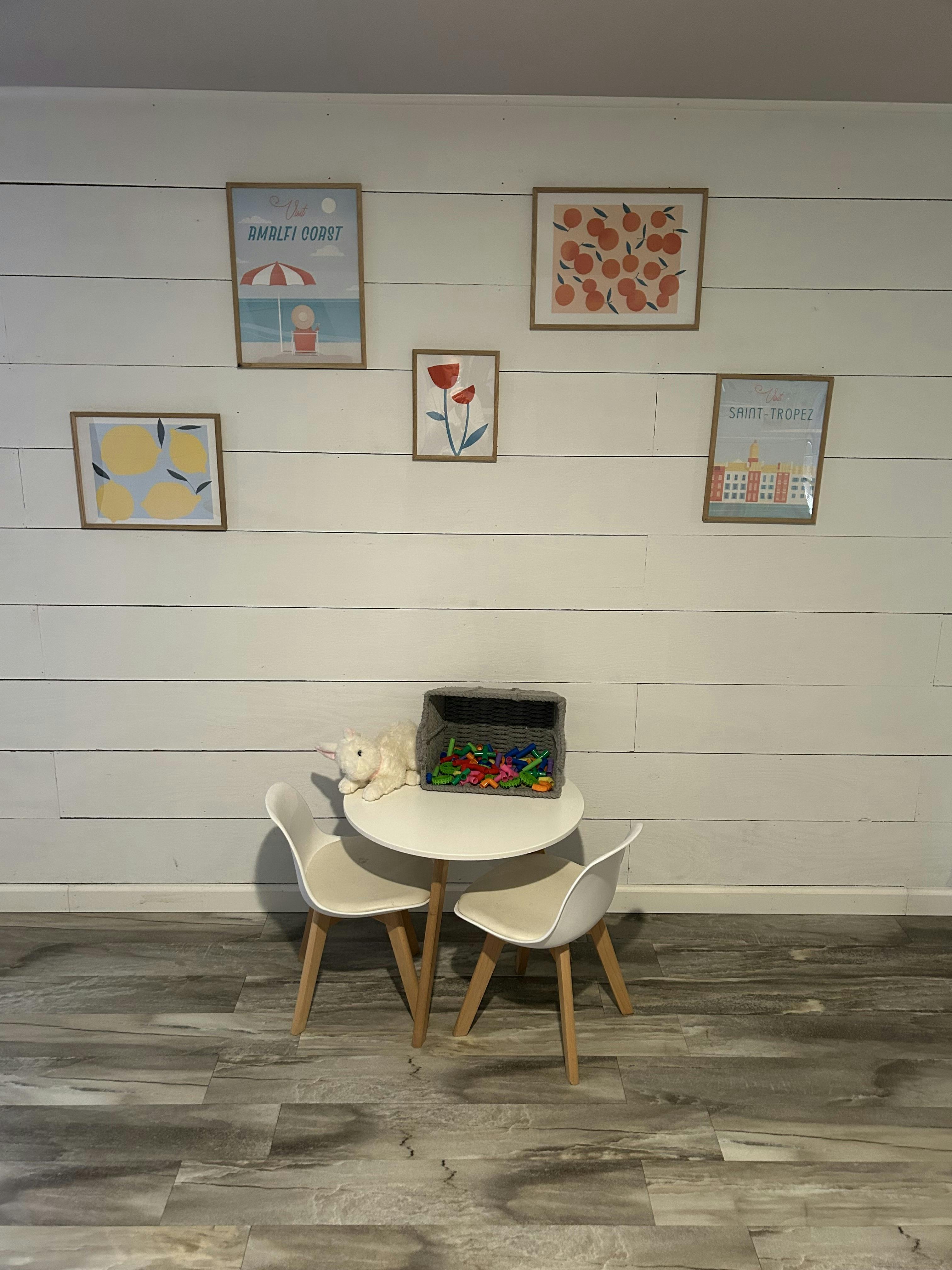 Small white table with two chairs, toy bin, and stuffed animal against a wall with framed art