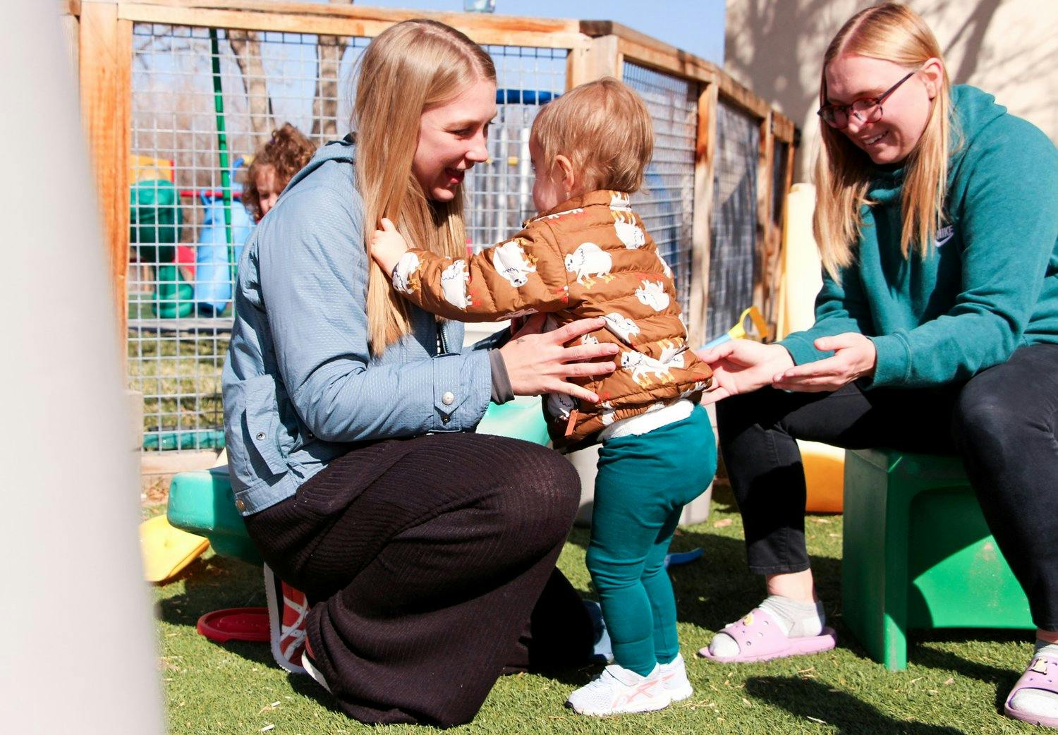Person helping a child stand outdoors while another person watches