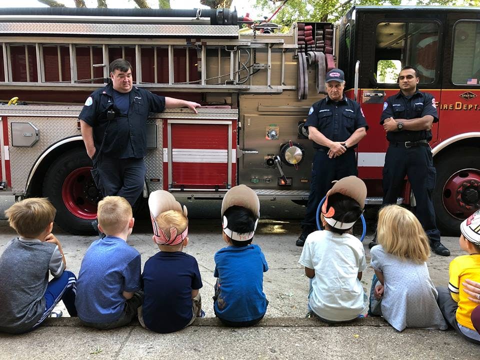 Children sitting on the ground in front of firefighters and a fire truck