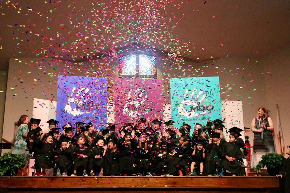 Children in graduation caps and gowns on a stage with confetti falling