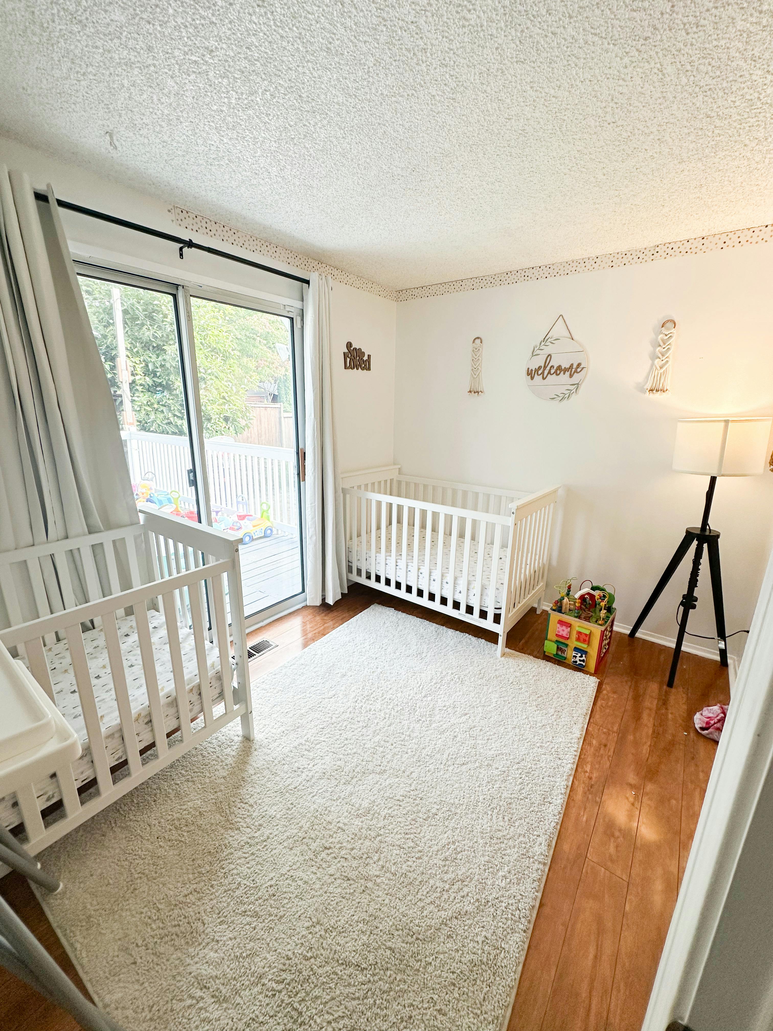 Two white cribs in a room with a sliding glass door and a white rug