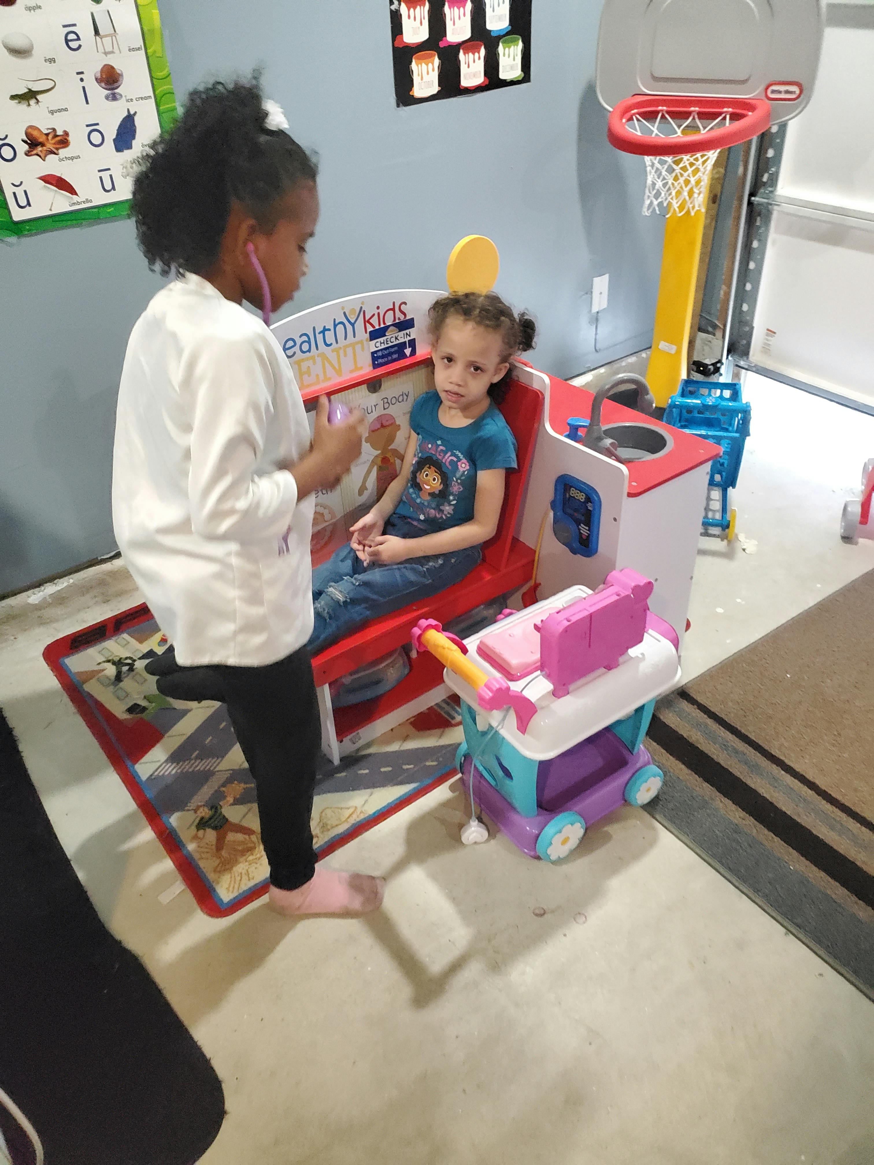Child in white coat with stethoscope examining another child sitting in a toy chair
