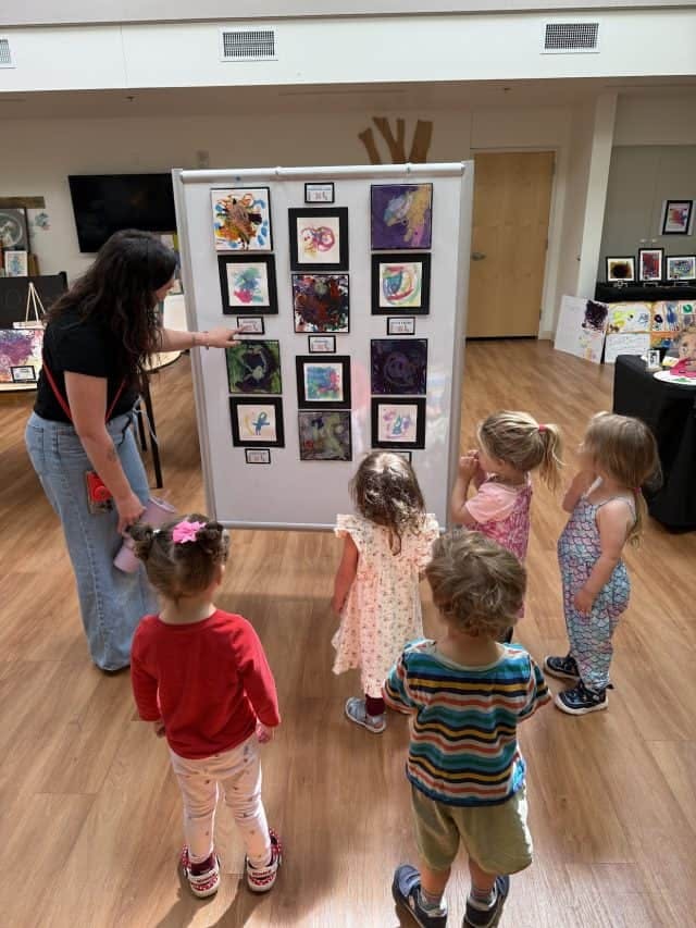 A person pointing at paintings on a display board while children look on