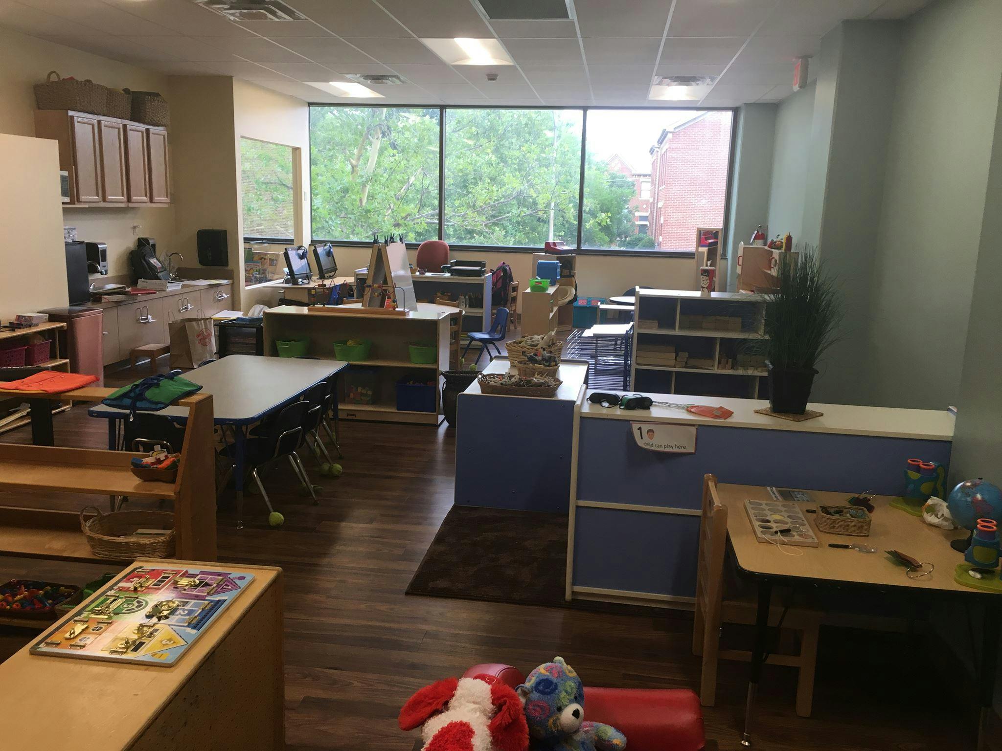 Classroom with tables, chairs, and shelves, large windows in background