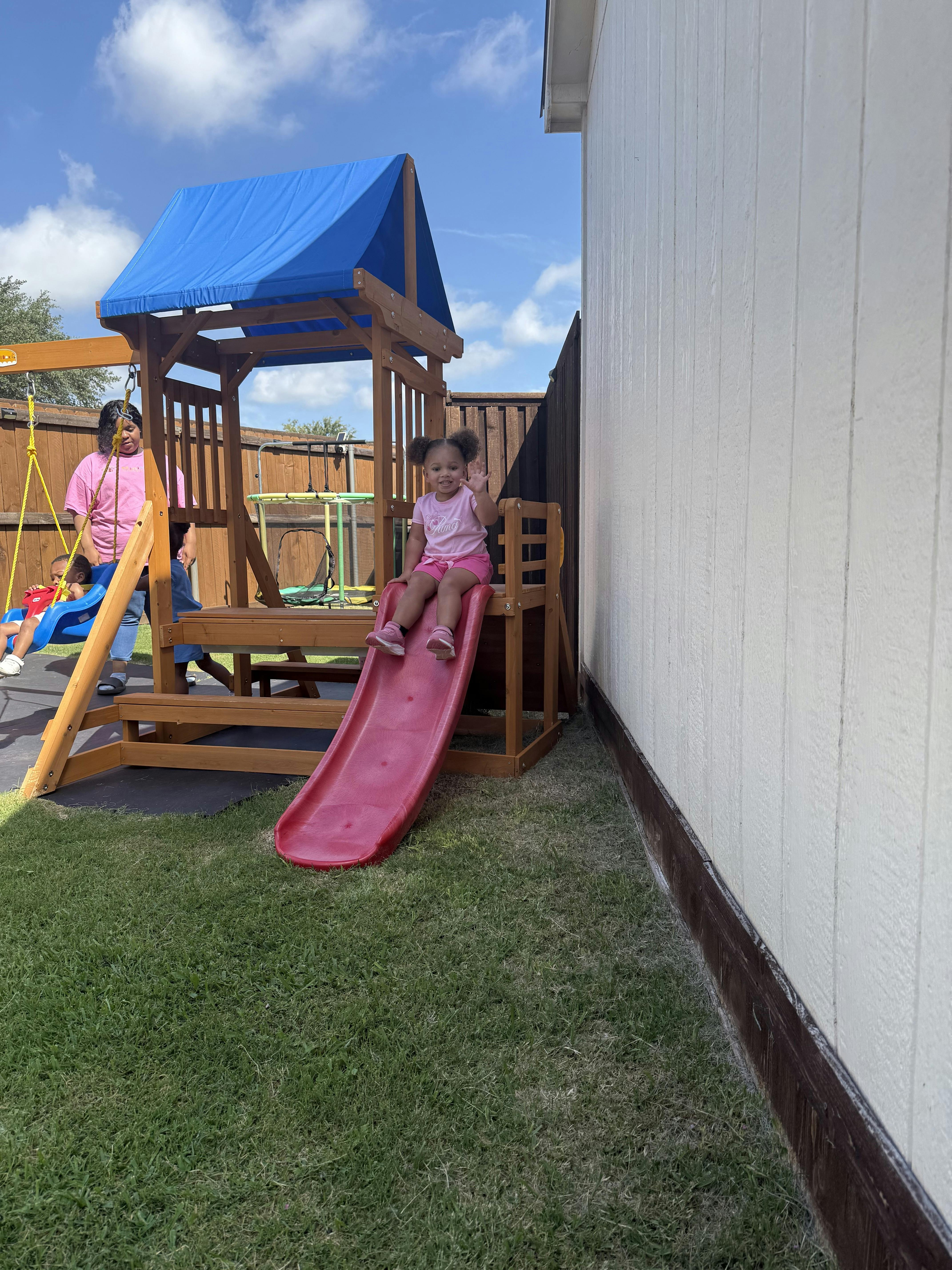Child waving while sitting at the top of a slide on a backyard playset