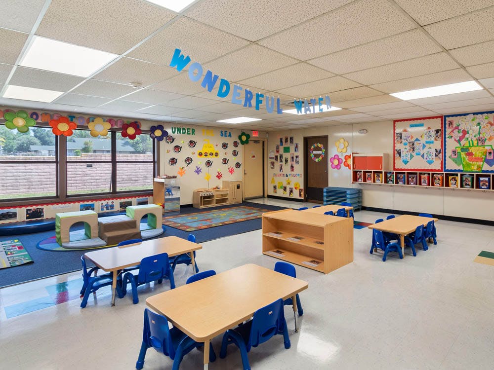 Empty classroom with small tables and chairs, colorful decorations, and large windows