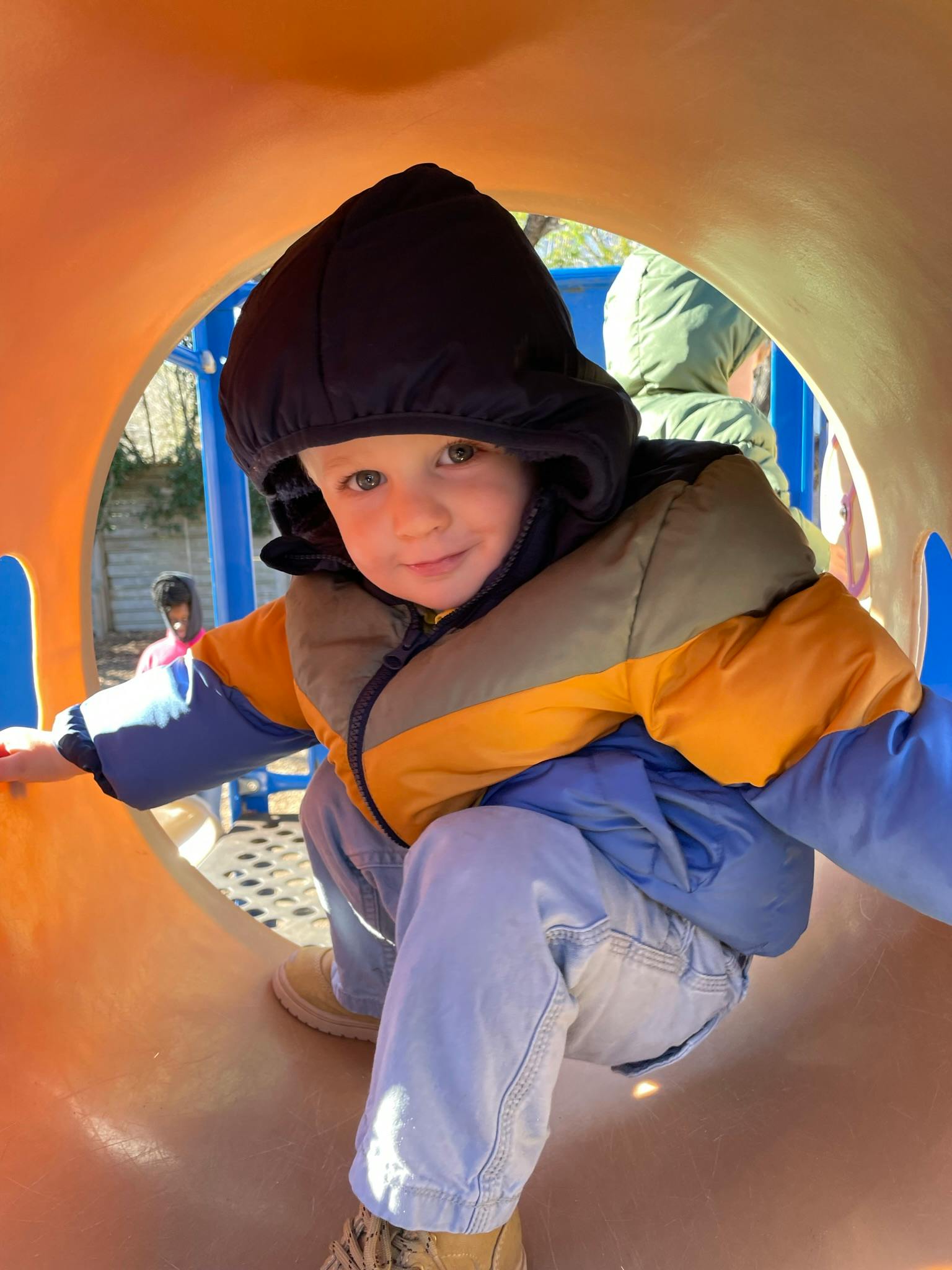 Child in a hooded jacket looking out from inside a playground tunnel