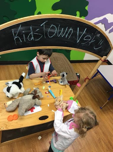 Two children playing with stuffed animals at a table under a "Kidstown Vet" sign