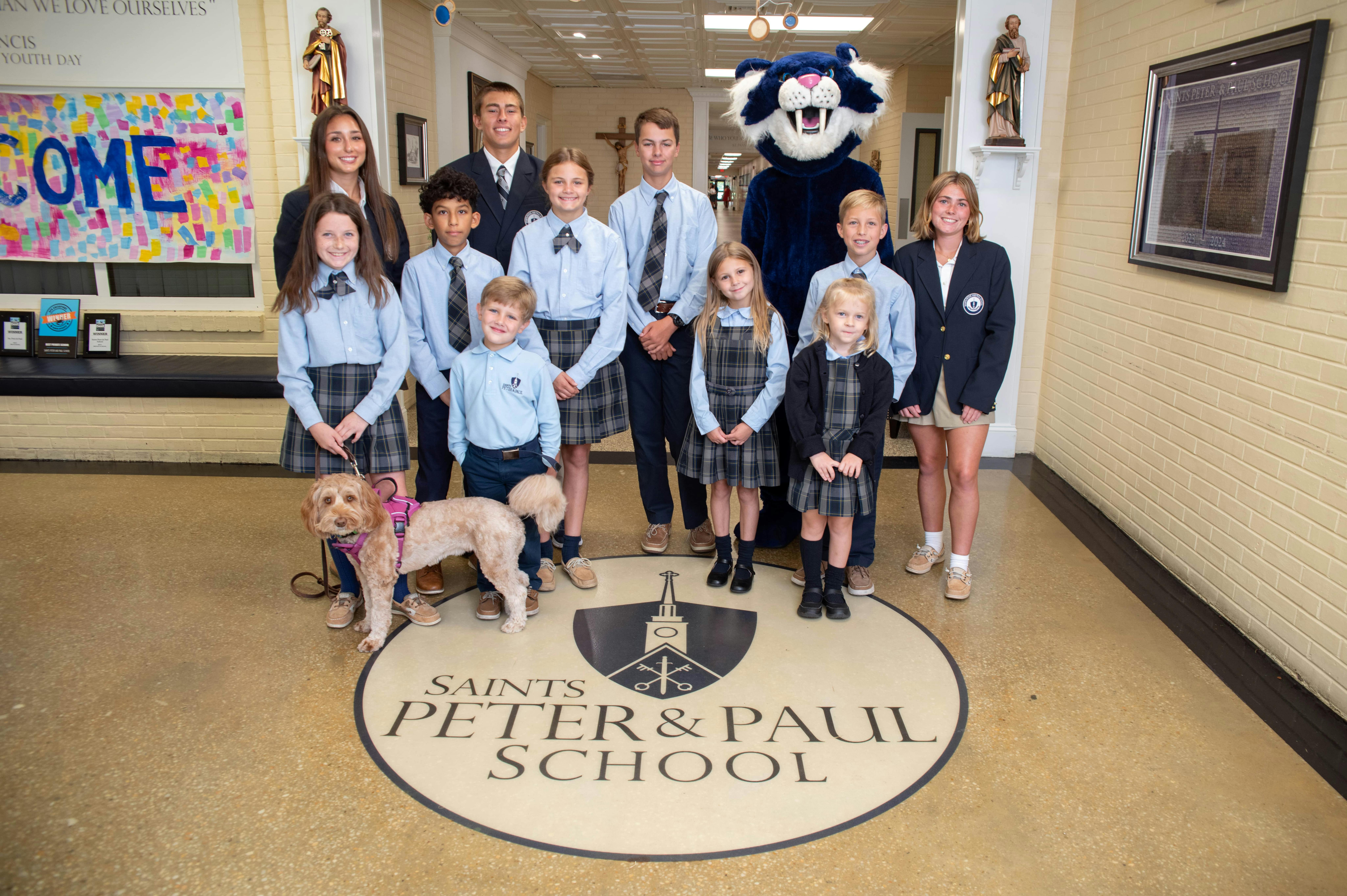 Group of adults, children, and a mascot standing on a school logo with a dog