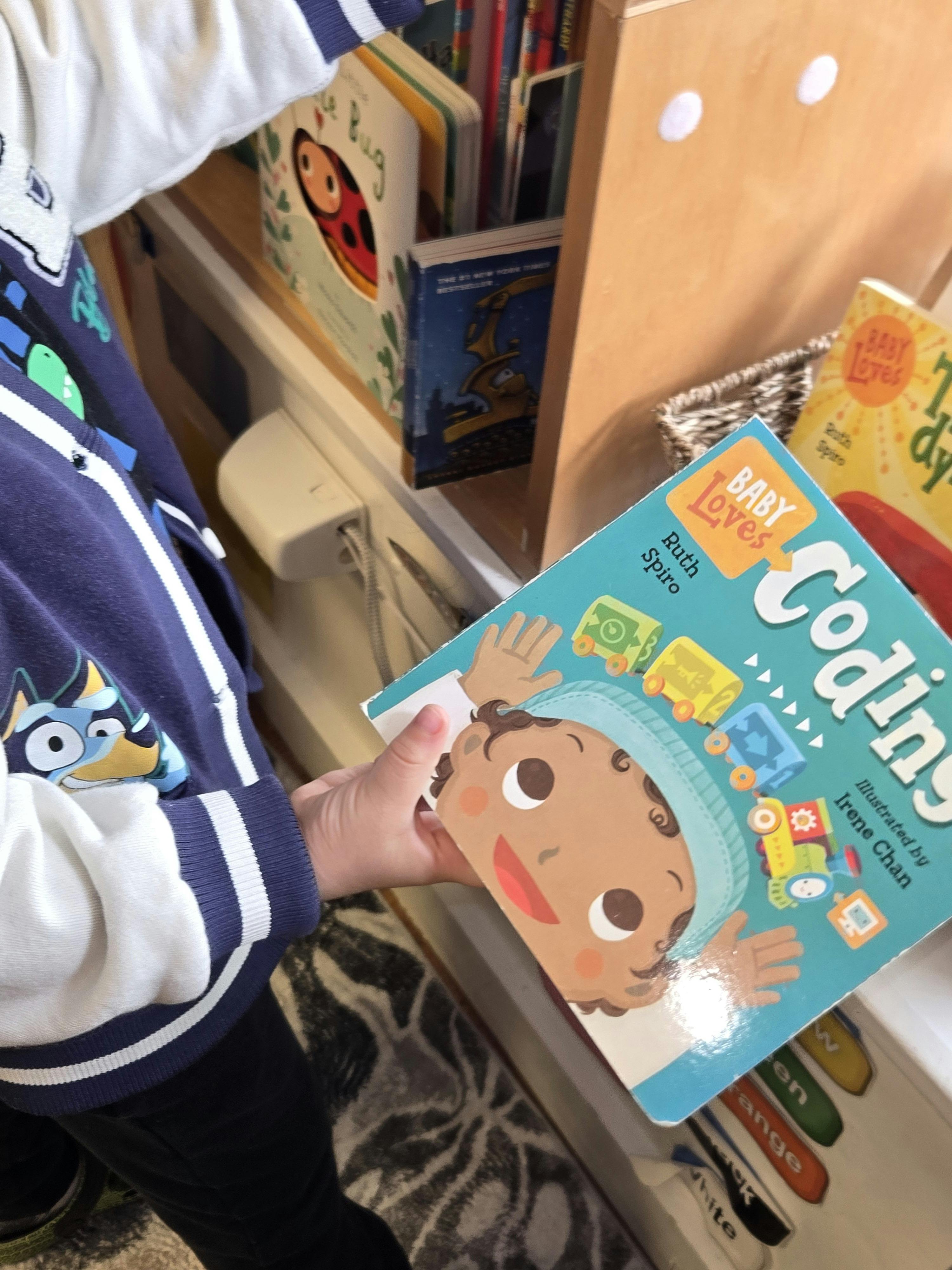 Child in a blue and white jacket holding a book titled "Baby Loves Coding"