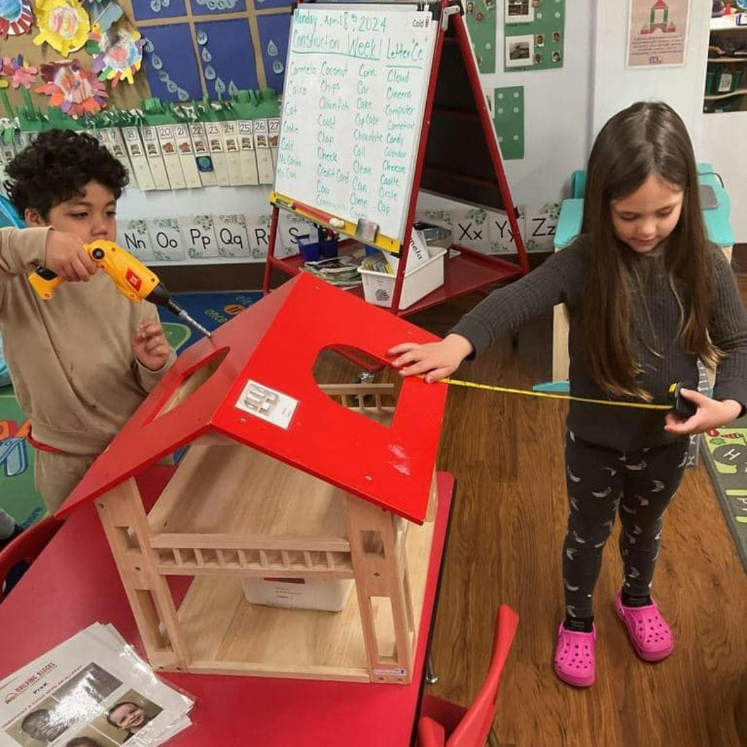 Two children playing with a dollhouse, one holding a drill, one measuring