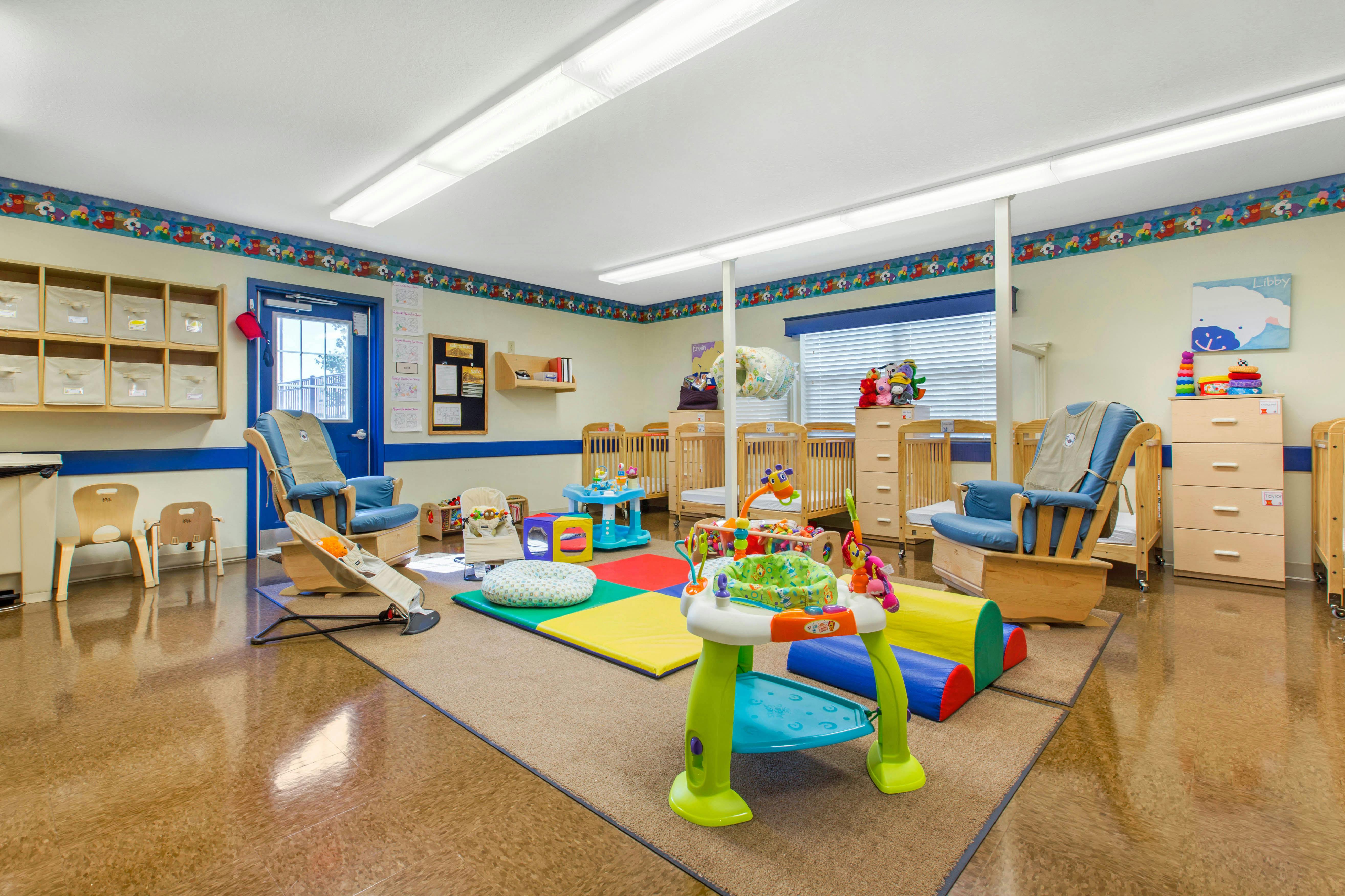 A childcare room with cribs, play mats, rocking chairs, and small furniture