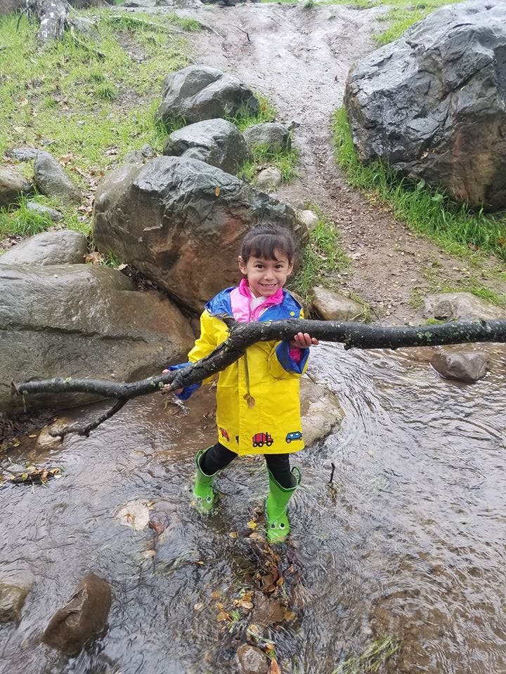 Child in a yellow raincoat and green boots holding a stick in a stream