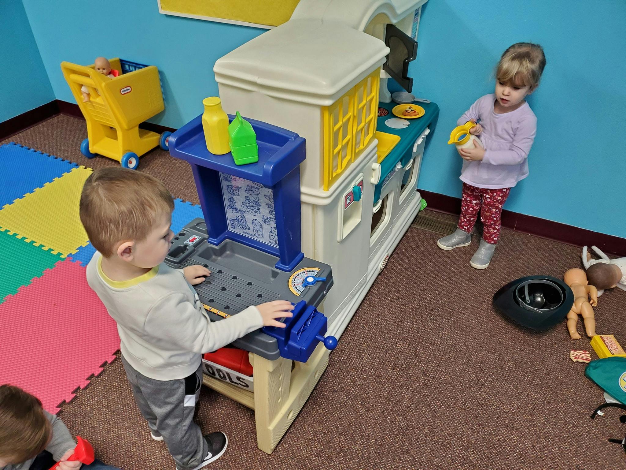 Child playing at a toy workbench, another child standing nearby