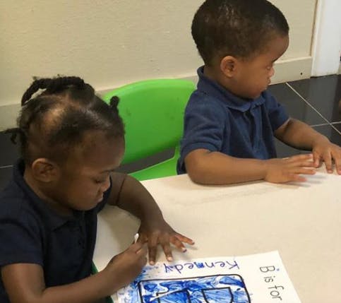 Two children sitting at a table, one coloring a paper with text "B is for"
