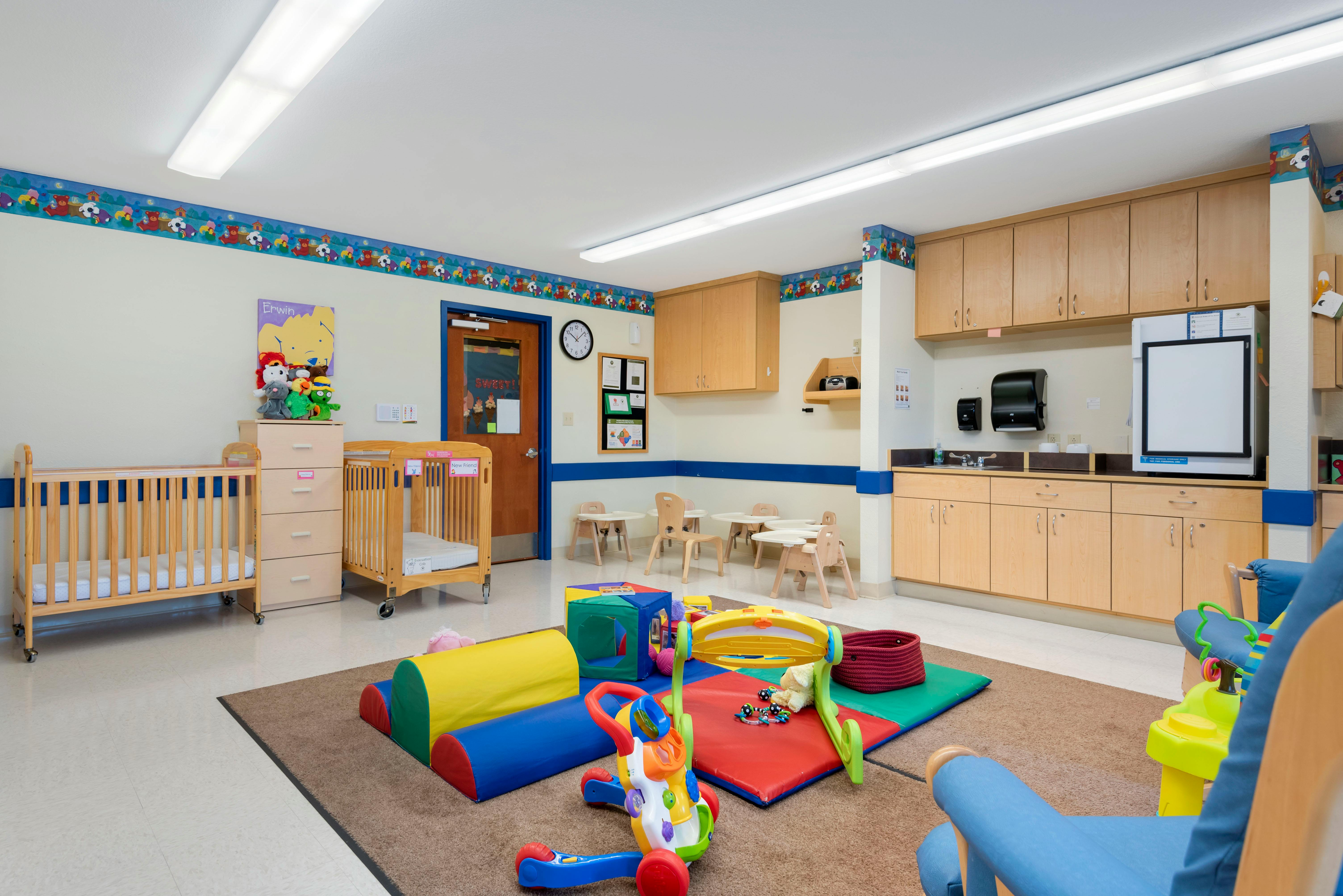 A colorful childcare classroom with cribs, play mats, and small tables